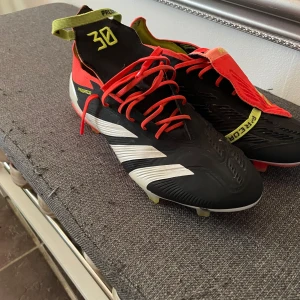 Adidas Predator fotbollsskor - Snygga Adidas Predator fotbollsskor i svart, röd och vit med snörning. Skorna har en modern design med tydliga logotyper och en slitstark sula för optimalt grepp på planen.