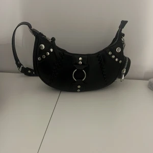 Svart axelväska med kedjedetaljer - Snygg svart axelväska i skinn med coola kedjedetaljer och nitar. Väskan har en dragkedja upptill och en justerbar axelrem med nitar. Perfekt för att ge din outfit en edgy touch.