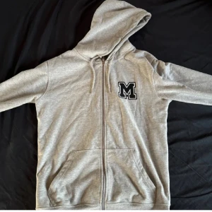 Grå hoodie med dragkedja och bokstavsmärke - Snygg grå hoodie med dragkedja och en stor bokstav 'M' på bröstet. Perfekt för en avslappnad stil med huva och fickor framtill.