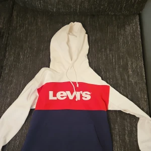 Flerfärgad hoodie från Levi's - Säljer en snygg flerfärgad hoodie från Levi's i storlek S. Tröjan har en vit huva och ärmar, röd mittsektion med Levi's-logga, och en mörkblå nederdel. Perfekt för en avslappnad stil.