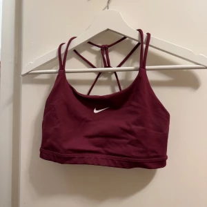 Vinröd sporttopp från Nike - Snygg vinröd sporttopp från Nike med smala axelband och korsad ryggdesign. Perfekt för träning med sin bekväma passform och stilrena look. Den har en liten vit Nike-logga framtill. Vet ej storlek för den är avklippta men gissar på M 