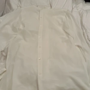 Vit blus från Patachou - Elegant vit blus från Patachou med långa ärmar och knappar framtill. Blusen har en stilren design med volangdetaljer vid halsen som ger en extra touch av elegans. Perfekt för speciella tillfällen eller en stilfull vardag.