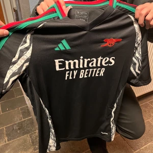 Svart Arsenal fotbollströja från Adidas - Snygg svart Arsenal fotbollströja från Adidas med korta ärmar och coola detaljer i vitt och grönt. Tröjan har 'Emirates Fly Better' tryckt på framsidan och 'Saka 7' på baksidan. Perfekt för alla Arsenal-fans! ⚽ helt ny också 