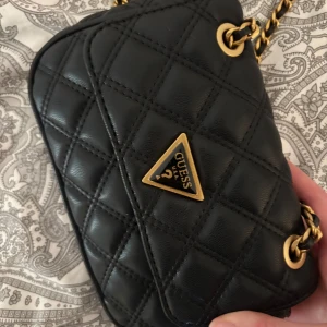 Svart quiltad axelväska från Guess - Snygg svart axelväska från Guess med quiltat mönster och guldfärgade kedjedetaljer. Väskan har en triangulär logotyp på framsidan och stängs med magnetknappar. Perfekt för att ge en elegant touch till din outfit.