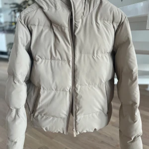 Beige pufferjacka från Zara - Snygg beige pufferjacka från Zara med dragkedja och huva. Perfekt för kyliga dagar med sin vadderade design. Jackan har en stilren look och är lätt att matcha med olika outfits. Storlek S