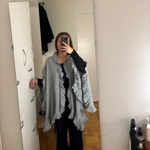 Grå poncho med volangdetaljer - Säljer en stilren grå poncho med volangdetaljer. Perfekt för lager-på-lager och ger en elegant touch till din outfit. Den är onesize och passar de flesta. Super fint skick, använd endast 2 gånger!