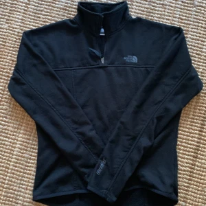 Svart fleece från The North Face - Säljer en svart fleece från The North Face i storlek S. Tröjan har en halv dragkedja och hög krage, perfekt för kyliga dagar. Tillverkad i 100% polyester för extra värme och komfort. En stilren och funktionell tröja för alla tillfällen.