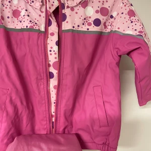 Rosa jacka med prickigt mönster - Säljer en söt rosa jacka med prickigt mönster i olika nyanser av rosa och lila. Jackan har en dragkedja framtill och är tillverkad i polyester. Perfekt för lekfulla dagar!