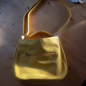 Gul axelväska - Snygg gul axelväska i syntetmaterial med justerbar axelrem. Väskan har en praktisk dragkedja framtill för enkel förvaring av småsaker. Perfekt för att lysa upp din outfit med en färgklick! Väskan har Inga täcken på användning. 
