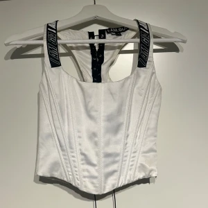 Vit korsett från I.AM.GIA - Snygg vit korsett från I.AM.GIA med svarta axelband som har logotypen tryckt. Den har en åtsittande passform och snörning i ryggen för justering. 