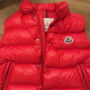 Röd dunväst från Moncler - Snygg röd dunväst från Moncler för barn storlek 10 år med dragkedja och knappar. Perfekt för kyliga dagar med sin quiltade design och hög krage. Västen har två praktiska fickor med dragkedja och Moncler-logga på bröstet. Storlek 10 år 