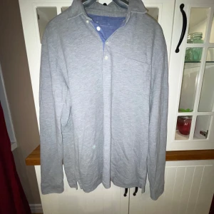 Grå långärmad pikétröja - Säljer en stilren grå långärmad pikétröja med knappar och bröstficka, från dressman. Som är i storlek xxl men sitter mer som en L/XL