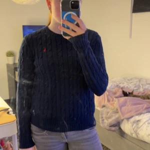 Mörkblå stickad tröja från Polo Ralph Lauren - Snygg mörkblå stickad tröja från Polo Ralph Lauren med kabelstickat mönster och röd logga på bröstet. Tröjan har långa ärmar och rund halsringning. Perfekt för kyliga dagar!