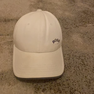 Vit keps från Hugo Boss - Snygg vit keps från Hugo Boss med broderad logga på framsidan. Perfekt för en stilren look. Justerbar passform för optimal komfort, den är köpt för 950kr och är knappt använd jätte fin till sommaren tveka inte på prisförslag!