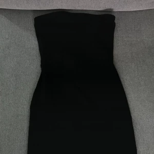 Svart axelbandslös klänning från Bershka - Snygg svart axelbandslös klänning från Bershka. Perfekt för en stilren look med sin enkla design och figurnära passform. Klänningen är kort och passar utmärkt för festliga tillfällen.