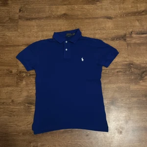Blå pikétröja från Ralph Lauren - Snygg blå pikétröja från Ralph Lauren i slim fit. Tröjan har en klassisk krage med knappar och det ikoniska broderade logotypen på bröstet. Perfekt för en stilren look.