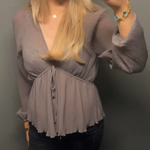  blus med knyt och volangdetaljer - Säljer en elegant grå blus med v-ringning och knyt framtill. Blusen har långa ärmar och fina volangdetaljer vid nederkanten. Perfekt för en stilren look. Är m men passar s 