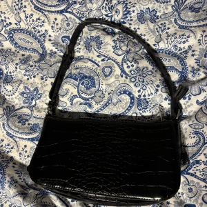 Svart axelväska med krokodilmönster - Snygg svart axelväska i syntet med krokodilmönster. Väskan har en justerbar axelrem. Använd några gånger men inga skador, jätte fin och bra kavalitet