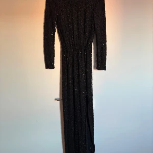 Svart glittrig långklänning - Elegant svart långklänning med glittrande detaljer och en djup v-ringning bak. Klänningen har långa ärmar och en slits som ger en sofistikerad look. Perfekt för festliga tillfällen. Storlek 34. 