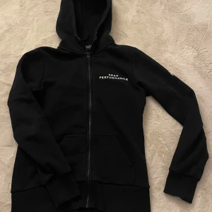 Svart hoodie från Peak Performance - Snygg svart hoodie från Peak Performance med dragkedja och huva. Perfekt för en avslappnad stil. Den har en enkel design med logotypen broderad på bröstet. Den är i storlek M men den är liten i storlek så passar S.