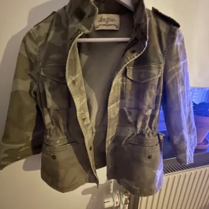 Camouflagejacka från Zara Woman - Snygg camouflagejacka från Zara Woman med dragkedja och knappar. Jackan har en normal passform och är perfekt för höst och vår. Den har lite kortare ärmar och praktiska fickor framtill.