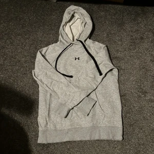 Under armour hoodie - Säljer en grå hoodie från Under Armour med svart logga på bröstet. Den har en justerbar huva med svarta snören och en stor magficka. Perfekt för en avslappnad stil. skick 8/10! Storlek M, skulle säga att den passar cirka 170cm. Tveka inte med att ställa frågor!