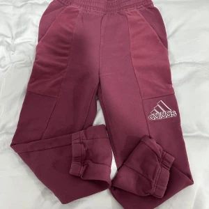 Vinröda mjukisbyxor från Adidas - Säljer ett par vinröda mjukisbyxor från Adidas med elastisk midja och snörning. Byxorna har en broderad Adidas-logga på benet och är tillverkade i mjukt material för extra komfort.