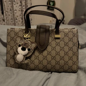 Brun mönstrad handväska med nalleberlock - Snygg handväska med klassiskt mönster och en söt nalleberlock. Väskan har bruna handtag och en avtagbar axelrem i grönt och rött. Perfekt för att ge din outfit en lyxig touch.