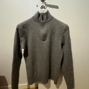 Ralph Lauren Cashmere - Snygg grå tröja från Ralph Lauren med halv dragkedja och broderad logga på bröstet. Perfekt för kyligare dagar med sin mjuka och bekväma passform.