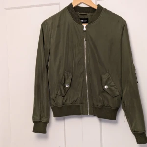 Olivgrön bomberjacka från FB Sister - Snygg olivgrön bomberjacka från FB Sister med dragkedja framtill och ribbade muddar. Jackan har två fickor med knappar och en klassisk bomberstil. Perfekt för en cool och avslappnad look.