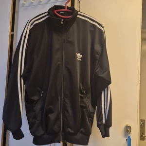 Svart träningsjacka från Adidas - Snygg svart träningsjacka från Adidas med klassiska vita ränder längs ärmarna. Jackan har dragkedja framtill och två fickor med dragkedjor. Perfekt för sportiga aktiviteter eller som en stilren vardagsjacka.