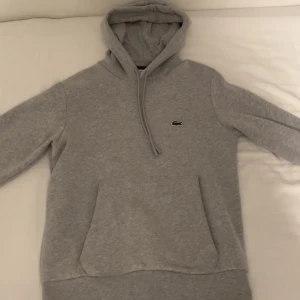 Lacoste hoodie  - Säljer en stilren grå hoodie från Lacoste helt ny, med klassisk krokodillogga på bröstet. Tröjan har en bekväm passform samt justerbar huva. Perfekt för en avslappnad look. Storlek XS men passar storlek S.
