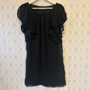 Svart klänning från Saint Tropez - Elegant svart klänning från Saint Tropez med volangdetaljer på ärmarna. Den har en luftig passform och är perfekt för en stilren look. 