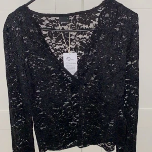 Svart spetsblus från Gina Tricot - Snygg svart spetsblus från Gina Tricot med v-ringning. Perfekt för en elegant look. Blusen har långa ärmar och ett genombrutet mönster som ger en stilren touch.