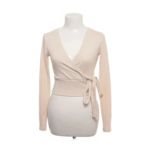 Beige omlottopp från NA-KD - Säljer en stilren beige omlottopp från NA-KD i storlek XS. Toppen har långa ärmar och ett knytband i midjan för en smickrande passform. Perfekt för både vardag och festliga tillfällen.