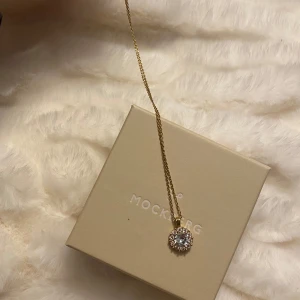 Lily and rose halsband - Ett jättefint guldigt halsband från lily and rose! Med utseende av en blomma mer än vad deras vanliga design brukar se ut. Små vita stenar längs stenen i mitten som även är ljusblå, som ja försöker visa på bild 3. Jättefint skick. 