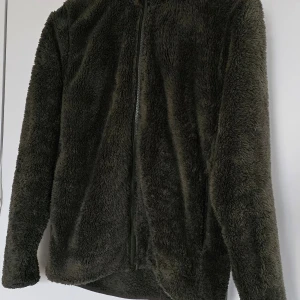 Grön teddyjacka - Mysig grön teddyjacka med dragkedja framtill. Perfekt för kyliga dagar med sin mjuka och varma känsla. Jackan har långa ärmar och en enkel design som passar till det mesta.