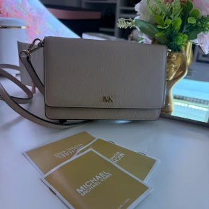 Beige axelväska från Michael Kors - Snygg beige axelväska från Michael Kors i skinn. Väskan har en elegant design med ett guldfärgat MK-emblem på framsidan. Den har en justerbar axelrem och flera fack inuti för kort och småsaker. Perfekt för att hålla ordning på dina tillhörigheter med stil.