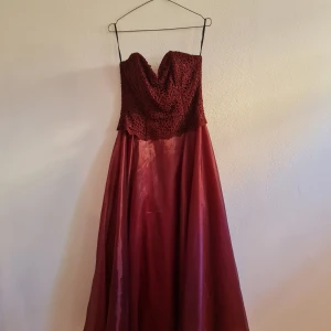 Vinröd långklänning med spetsdetaljer - Säljer en superfin vinröd långklänning med spetsdetaljer på överdelen. Klänningen är axelbandslös och har en vacker snörning på baksidan. Kjolen är i ett glansigt material som ger en lyxig känsla. Perfekt för bal eller fest! 💃 färgen är lite annorlunda ön på bilden