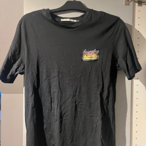 Svart t-shirt med tryck - Säljer en svart t-shirt med ett coolt tryck på bröstet. T-shirten är i bra skick och passar perfekt till vardags eller fest. Den är gjord av bomull och har en normal passform. Perfekt för dig som vill ha en enkel men stilren look!