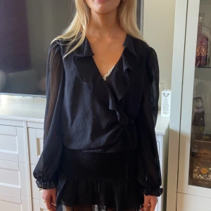 Svart blus med volangdetaljer - Säljer en elegant svart blus med volangdetaljer. Blusen har långa ärmar och en v-ringad hals. Materialet är lätt och luftigt, perfekt för både vardag och fest. Den har en knytning i midjan som ger en fin passform. Passar bra till både kjol och byxor.