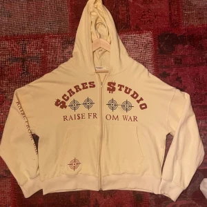Beige hoodie från Scares Studio - Säljer en beige hoodie från Scares Studio med texten 'RAISE FROM WAR' och måltavlor tryckta i rött på framsidan. Hoodien har en dragkedja framtill och långa ärmar. den har croped pass form. Perfekt för en avslappnad och cool stil. Köp direkt eller ge prisförslag 💛