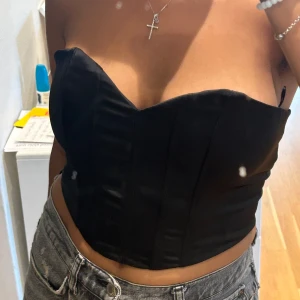 Svart korsett topp - Säljer en snygg svart korsett topp. Den är axelbandslös och har en tight passform som framhäver figuren. Perfekt för fest eller en utekväll. Toppen är i nyskick och har en stilren design som passar till många olika outfits. Dragkedja vid sidan så det blir enkelt att sätta på och av, dragkedjan är inte synlig. 