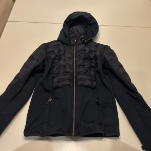 Mörkblå jacka från JH Collection - Säljer en snygg marinblå ridjacka från JH Collection i storlek XS. Jackan har en huva som går att ta av och dragkedja framtill. Den är quiltad på framsidan och baksidan samt på ärmarna. Perfekt för stallet!🐎