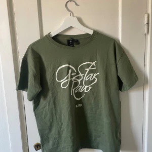 Grön t-shirt från G-Star RAW - Säljer en snygg grön t-shirt från G-Star RAW. Den är i storlek L och har en cool logga på framsidan. T-shirten är gjord av bomull och är superbekväm att ha på sig. Perfekt för vardagsbruk eller en avslappnad dag ute. Den är i nyskick med tagg och har aldrig använts.