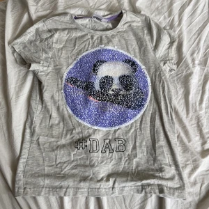 Grå t-shirt med panda #DAB från emoji - Säljer en grå t-shirt från emoji med en cool panda som dabbar på framsidan. T-shirten har korta ärmar och är gjord av mjukt bomullsmaterial. Perfekt för vardagsbruk eller för att visa upp din dab-skills! 🐼✨