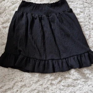 Svart kjol med volang - Säljer en superfin svart kjol med volangdetaljer samt fickor på båda sidorna. Kjolen är kort och har en elastisk midja för en bekväm passform. Perfekt för både vardag och fest! 🖤