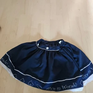 Mörkblå kjol med Alice in Wonderland-tema - Säljer en superfin mörkblå kjol med Alice in Wonderland-tema. Kjolen har en vit kant längst ner och texten 'Alice in Wonderland' broderad i vitt. Den har en söt vit knappdetalj i midjan och är perfekt för alla som älskar sagor och magi. Passar perfekt till fest eller cosplay!