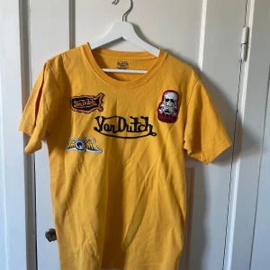 Gul t-shirt från Von Dutch - Säljer en cool gul t-shirt från Von Dutch. Den har flera broderade detaljer, inklusive en dödskalle och vingar. T-shirten är i bomull och har en normal passform. Perfekt för en avslappnad stil!