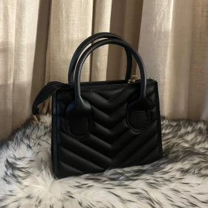 Svart quiltad handväska - Säljer en snygg liten svart quiltad handväska i skinn. Den har två handtag, en dragkedja och en avtagbar axelrem. Perfekt för både vardag och fest! Aldrig använd